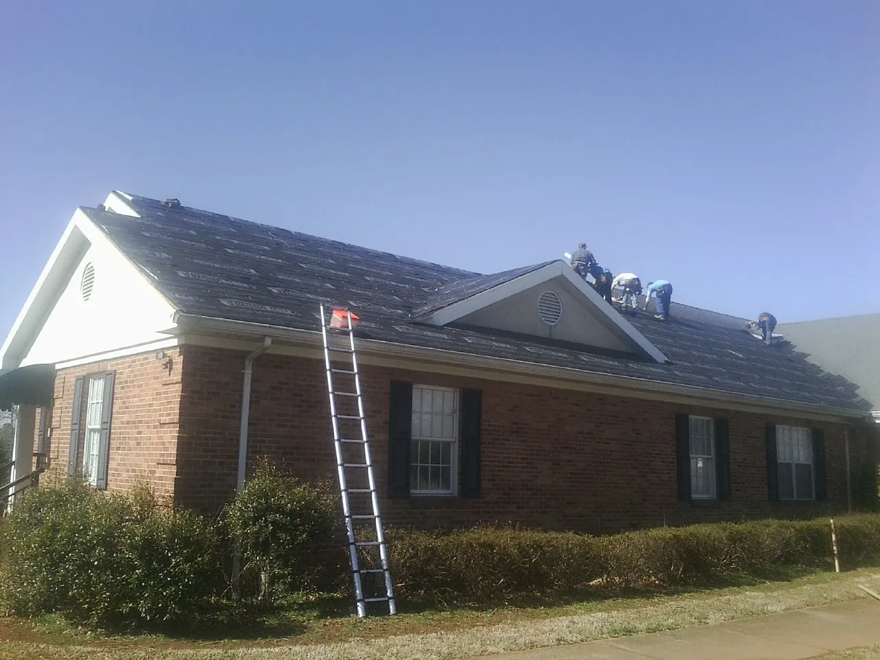 Crew tearing shingles off a brick ranch roof with a ladder set — Emergency Roof Repair work by our crew in Twin Lakes